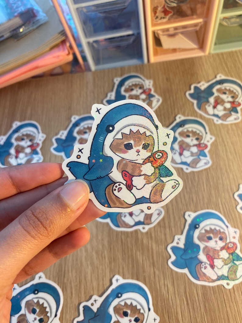 May include: A die-cut sticker featuring a brown tabby cat wearing a blue shark costume. The cat is holding a small, colourful fish. The sticker has a holographic finish with small star accents and black 'x' and 'o' symbols printed on the sides.