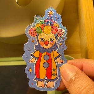 May include: A holographic sticker featuring a cartoon clown character with a red nose, wearing a colorful polka-dot hat and a yellow and red striped jumpsuit with blue ruffles. The background is a deep blue with small yellow star accents.