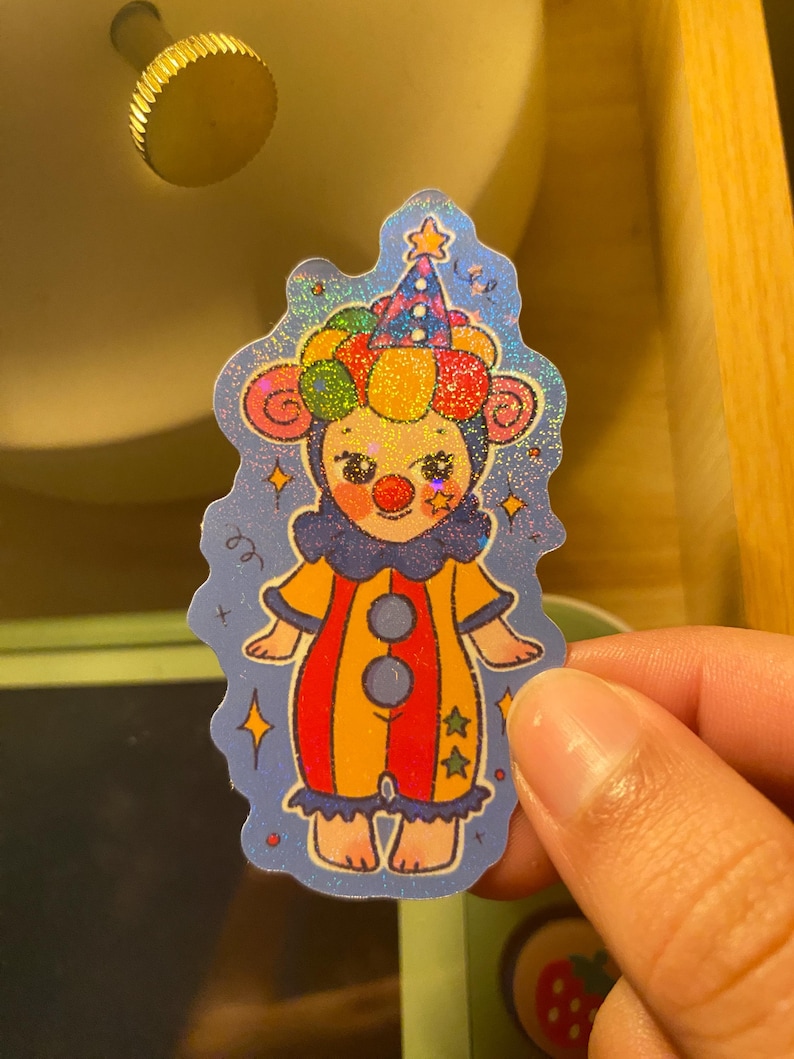 May include: A holographic sticker featuring a cartoon clown character with a red nose, wearing a colorful polka-dot hat and a yellow and red striped jumpsuit with blue ruffles. The background is a deep blue with small yellow star accents.