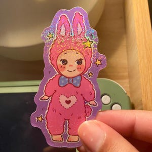 May include: A holographic sticker featuring a cartoon character wearing a pink bunny onesie with a blue bow tie and a heart-shaped patch on the chest. The character has rosy cheeks and is surrounded by small yellow stars against a purple background.
