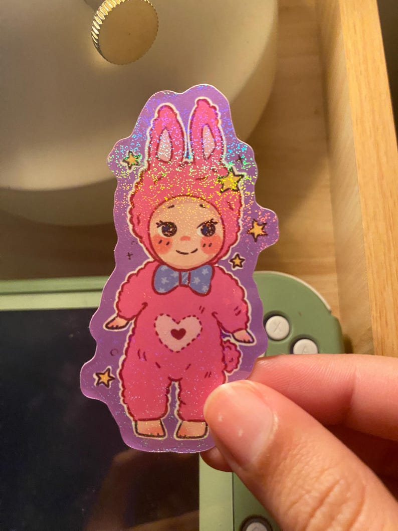 May include: A holographic sticker featuring a cartoon character wearing a pink bunny onesie with a blue bow tie and a heart-shaped patch on the chest. The character has rosy cheeks and is surrounded by small yellow stars against a purple background.
