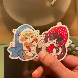 May include: Two holographic stickers featuring illustrated cats. One sticker shows an orange cat wearing a blue shark costume holding a fish. The other sticker shows a dark-furred cat wearing a red strawberry hood while holding a strawberry.