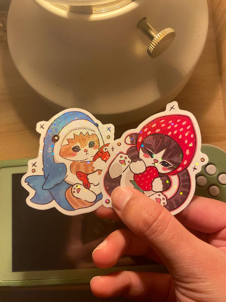 May include: Two holographic stickers featuring illustrated cats. One sticker shows an orange cat wearing a blue shark costume holding a fish. The other sticker shows a dark-furred cat wearing a red strawberry hood while holding a strawberry.