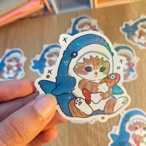 May include: A die-cut sticker featuring a brown and white cat wearing a blue shark costume. The cat is holding a small, colourful fish toy. The sticker has a holographic finish with small star-shaped sparkles and includes small black x symbols.