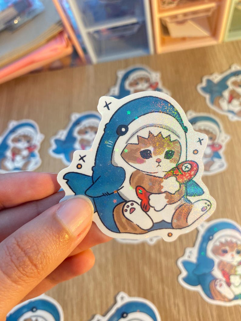 May include: A die-cut sticker featuring a brown and white cat wearing a blue shark costume. The cat is holding a small, colourful fish toy. The sticker has a holographic finish with small star-shaped sparkles and includes small black x symbols.