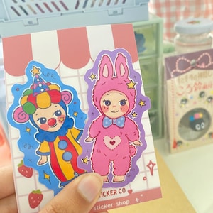 May include: Two die-cut stickers on a pink and white grid backing card. One sticker features a character in a colorful clown costume with a hat. The other sticker shows a character in a pink bunny onesie with a blue bow tie and heart detail.