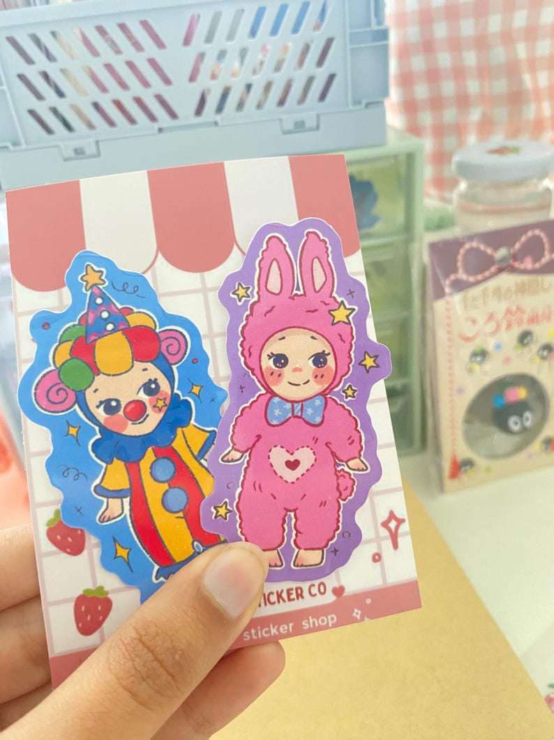 May include: Two die-cut stickers on a pink and white grid backing card. One sticker features a character in a colorful clown costume with a hat. The other sticker shows a character in a pink bunny onesie with a blue bow tie and heart detail.