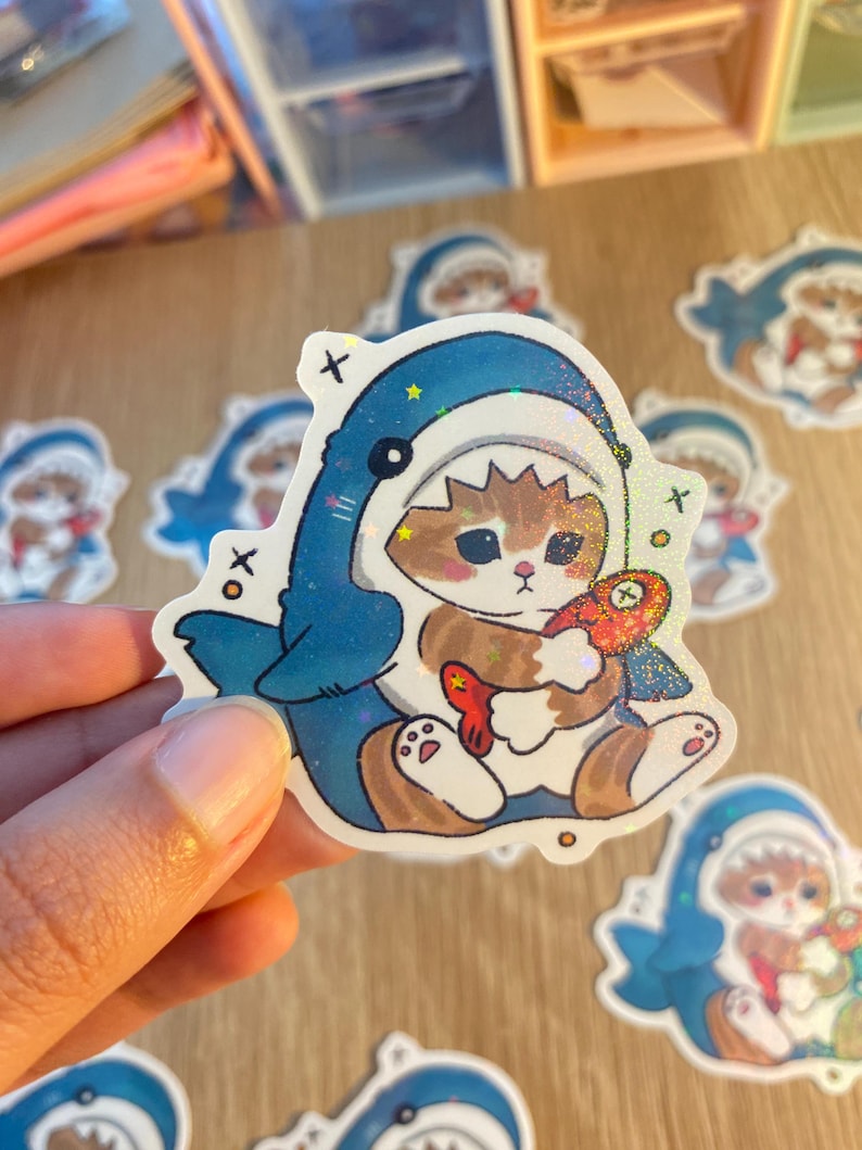 May include: A die-cut sticker featuring a brown tabby kitten wearing a blue shark costume with a jagged hood. The kitten holds a small orange fish with a cross-stitched eye. The sticker has a holographic finish with scattered star accents.