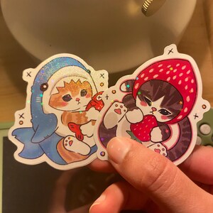 May include: Two holographic stickers featuring illustrated cats. One sticker shows an orange tabby cat wearing a blue shark costume holding a red fish. The other sticker shows a grey tabby cat wearing a red strawberry hood holding a strawberry.