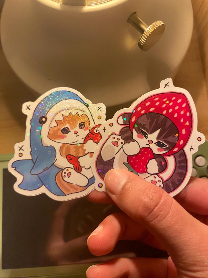 May include: Two holographic stickers featuring illustrated cats. One sticker shows an orange tabby cat wearing a blue shark costume holding a red fish. The other sticker shows a grey tabby cat wearing a red strawberry hood holding a strawberry.