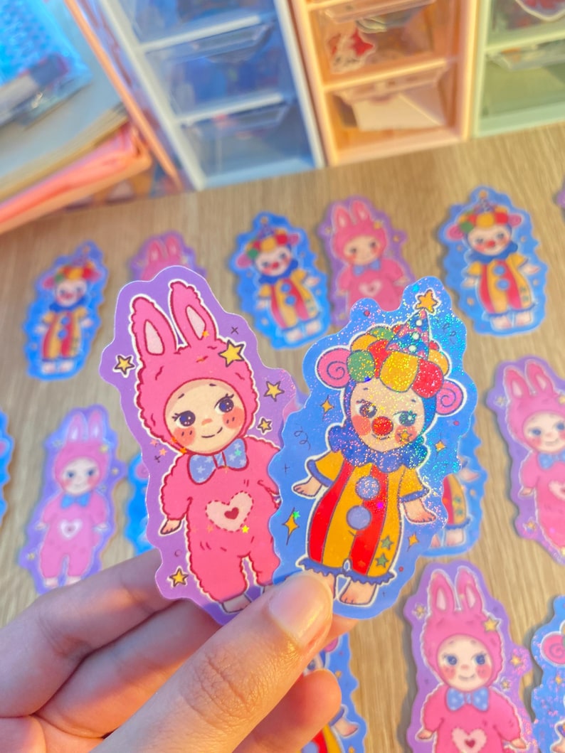 May include: Two die-cut stickers featuring cartoon characters. One is a pink rabbit-costumed figure with a blue bow tie and heart detail. The other is a clown-costumed figure in a yellow and red striped suit with a colorful hat and glitter finish.