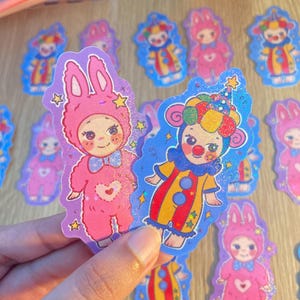 May include: Two glittery die-cut stickers featuring cartoon characters. One is a pink rabbit-costumed figure with a heart on its chest and a blue bow tie. The other is a clown-costumed figure with a colorful hat, red nose, and yellow and red suit.