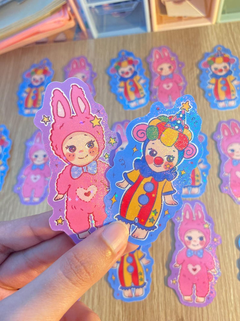 May include: Two glittery die-cut stickers featuring cartoon characters. One is a pink rabbit-costumed figure with a heart on its chest and a blue bow tie. The other is a clown-costumed figure with a colorful hat, red nose, and yellow and red suit.