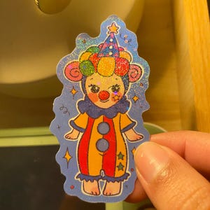 May include: A holographic sticker featuring a cartoon clown character with a red nose, wearing a colorful polka-dot hat and a yellow and red striped jumpsuit with blue ruffles. The background is a deep blue with small yellow star accents.