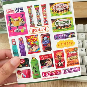 Könnte beinhalten: Ein Blatt mit bunten Aufklebern mit japanischen Snacks und Getränken. Die Aufkleber zeigen Bilder von beliebten Marken wie Pocky, Hello Panda und Kinoko no Yama. Die Aufkleber sind auf weißem Papier mit grünem Rand gedruckt. Der Text auf den Aufklebern ist in Japanisch.