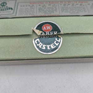 Op de afbeelding: Een close-up van een vintage groene doos met het A.W. Faber Castell logo. Het logo is een cirkelvormig embleem met de woorden "A.W. Faber" en "Castell" in witte letters. De doos heeft een lichtgroene kleur en een papieren label.