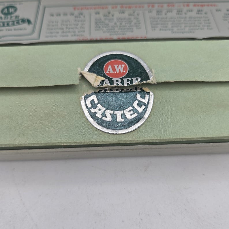 Op de afbeelding: Een close-up van een vintage groene doos met het A.W. Faber Castell logo. Het logo is een cirkelvormig embleem met de woorden "A.W. Faber" en "Castell" in witte letters. De doos heeft een lichtgroene kleur en een papieren label.