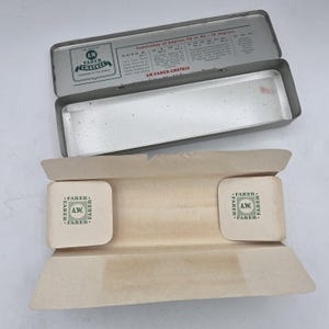 Op de afbeelding: Een open, vintage zilveren metalen etui met een papieren inzetstuk. De deksel heeft tekst waaronder "AW FABER-CASTECC". Het papieren inzetstuk heeft twee vierkante pads met het "FABER"-logo.