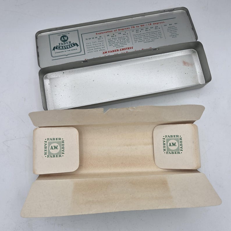 Op de afbeelding: Een open, vintage zilveren metalen etui met een papieren inzetstuk. De deksel heeft tekst waaronder "AW FABER-CASTECC". Het papieren inzetstuk heeft twee vierkante pads met het "FABER"-logo.