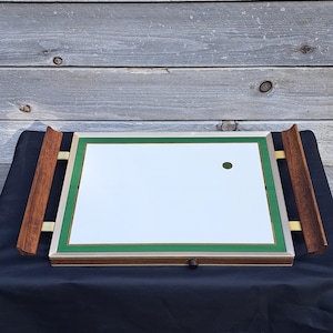 May include: Rectangular mirror with a green border and wooden handles. The mirror has a silver frame and a small hole near the top. The handles are made of dark wood and are attached to the sides of the mirror with brass-colored metal.