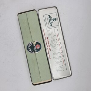 Op de afbeelding: Een open, vintage potloodkoker van Faber-Castell, in lichtgroen en zilver metaal. De koker is rechthoekig en bevat een papieren inzetstuk met tekst. Het Faber-Castell-logo staat op de koker.