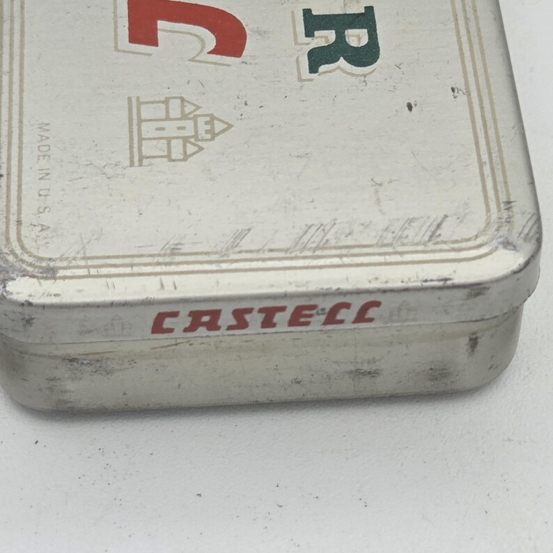 Op de afbeelding: Een rechthoekige, zilverkleurige metalen doos met het woord "CASTELL" in rode letters op de zijkant. De bovenkant van de doos heeft de letter "R" in groen en een kasteelillustratie. De woorden "MADE IN U.S.A." staan op de zijkant.