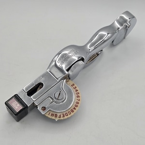 May include: A vintage silver Dymo-Mite tapewriter. The label maker has a handle and a dial with letters and numbers. The text "DYMO-MITE TAPEWRITER" is printed on the body. The model number is M-22, 1300W.