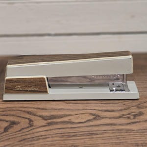 May include: A vintage Swingline stapler with a cream-colored body and a wood-grain top and accents. The stapler features the Swingline logo in a cursive font on the side. The stapler is on a wooden surface.