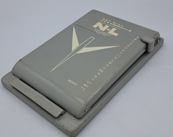May include: A gray vintage address book with the text "SET DIAL HERE" and "N-L" printed on the cover. The cover also features a white geometric design. The address book has an alphabetical dial and a memo section.