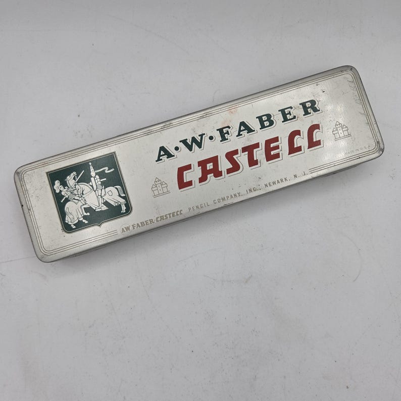 Op de afbeelding: Vintage A.W. Faber Castell etui. De rechthoekige metalen koffer is zilverkleurig met de woorden "A.W. Faber Castell" in groen en rood. Een groen embleem met een ridder te paard staat aan de linkerkant.