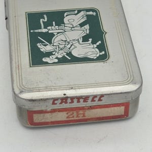 Op de afbeelding: Een rechthoekige metalen etui met een groen en wit embleem op het deksel. Het embleem toont twee figuren te paard. De onderkant van de etui heeft het woord "CASTELL" in rood en een label met "2H".