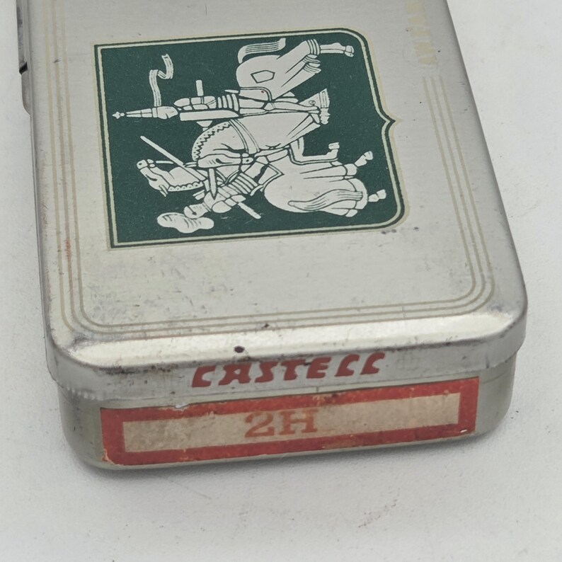 Op de afbeelding: Een rechthoekige metalen etui met een groen en wit embleem op het deksel. Het embleem toont twee figuren te paard. De onderkant van de etui heeft het woord "CASTELL" in rood en een label met "2H".