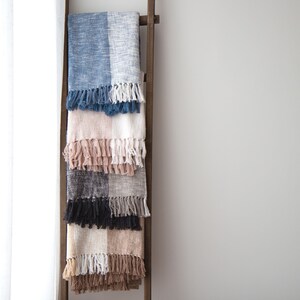 May include: A set of five woven blankets with fringe, stacked on a wooden ladder. The blankets are in shades of blue, pink, gray, black, and brown.