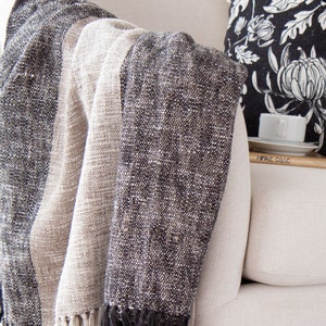 May include: A soft, striped throw blanket in shades of gray and beige, draped over a white armchair. The blanket has a textured weave and a fringe edge.