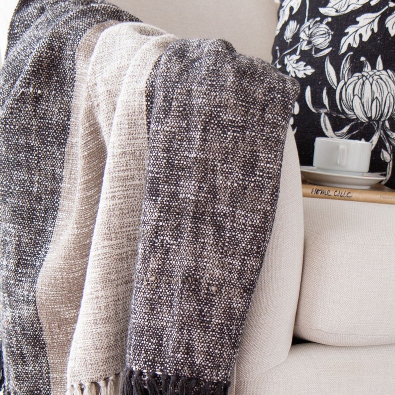 May include: A soft, striped throw blanket in shades of gray and beige, draped over a white armchair. The blanket has a textured weave and a fringe edge.
