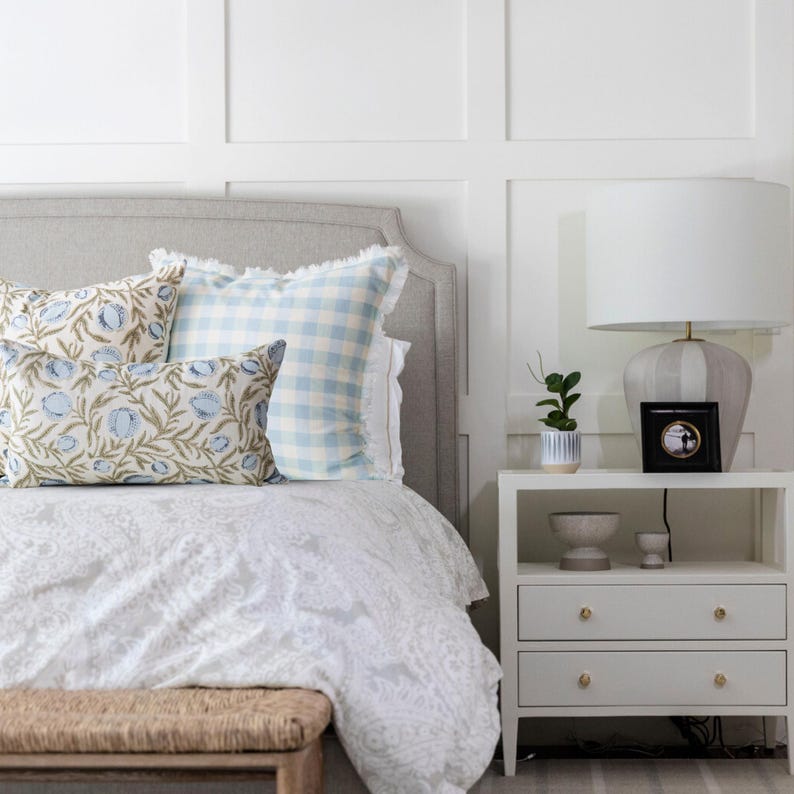 May include: A bedroom scene featuring a bed with decorative pillows in blue and white patterns. A white nightstand with gold knobs holds a lamp, a small plant, and a framed picture. The bed has a gray headboard and a white patterned comforter.
