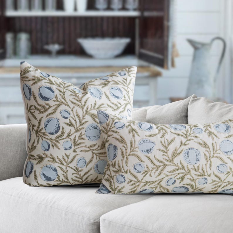 May include: Two decorative throw pillows with a botanical pattern. The pillows feature a cream-colored background with a repeating design of blue fruit and olive-green leaves. The pillows are placed on a white sofa.