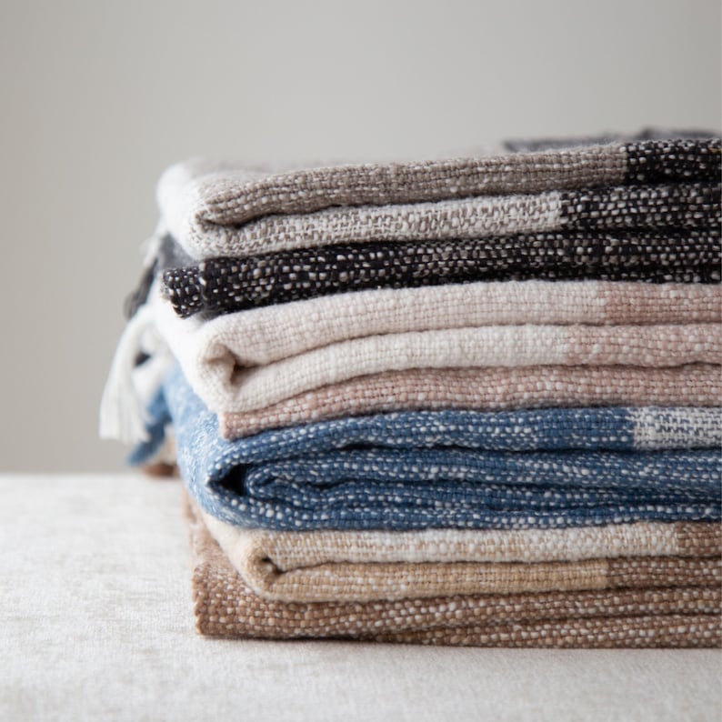 K&ouml;nnte beinhalten: Ein Stapel gewebter Decken in verschiedenen Farben, darunter Beige, Braun, Blau und Schwarz. Die Decken sind ordentlich gefaltet und haben eine strukturierte Oberfl&auml;che.