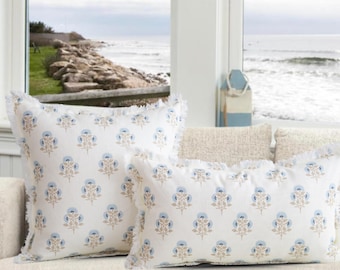 May include: Two white throw pillows with a blue and tan floral pattern and fringed edges. The pillows are rectangular and square, resting on a cream-colored sofa. The background shows a coastal view with the ocean and a rocky shoreline.