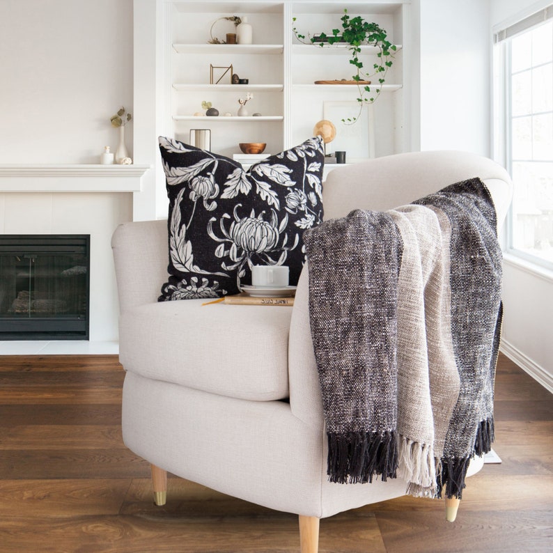 May include: A white armchair with a black and white floral throw pillow and a gray and beige throw blanket. The chair is sitting in front of a fireplace and a bookshelf.