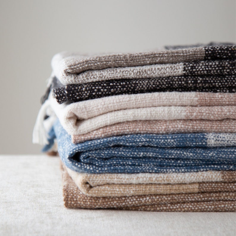 May include: A stack of woven blankets in various colors, including beige, brown, blue, and black. The blankets are folded neatly and have a textured surface.
