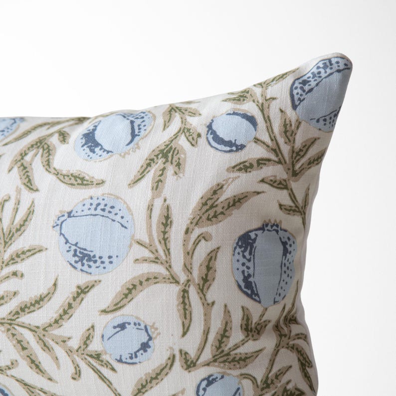 May include: A corner of a decorative pillow with a botanical pattern. The pillow features a cream-colored background with a repeating design of light blue fruit and olive-green leaves and stems.