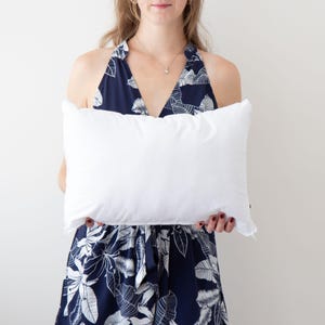 Puede incluir: Una almohada blanca con una funda blanca. La almohada está siendo sostenida por una persona que lleva un vestido floral azul y blanco.