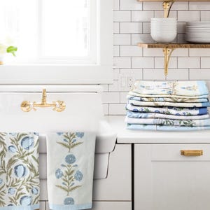 May include: A stack of folded kitchen towels with blue floral patterns and light blue borders sits on a white countertop. Two matching towels hang on a brass faucet. The towels feature botanical designs in blue, green, and white.