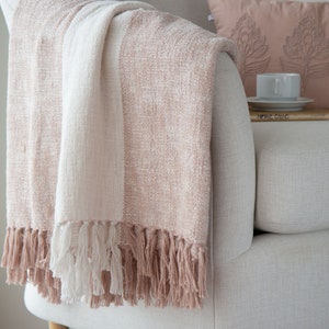 K&ouml;nnte beinhalten: Eine weiche, gestreifte Wolldecke in Wei&szlig; und Blush-Rosa. Die Decke hat einen Fransenrand und ist &uuml;ber einen wei&szlig;en Sessel gelegt.