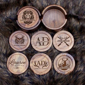 May include: A set of round wooden coasters with a matching storage holder. Each coaster features unique laser-engraved designs including a lion, a skull with roses, crossed rifles, a deer in a mountain landscape, and various personalised names and initials.