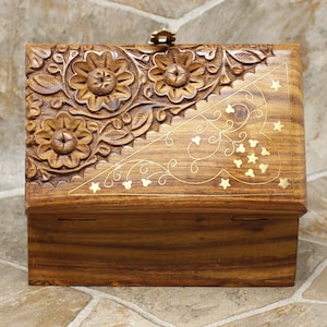 Puede incluir: Una caja de madera con intrincadas tallas florales y detalles de lat&oacute;n. La parte superior presenta flores y hojas talladas, con un cierre de lat&oacute;n. La caja es de color marr&oacute;n c&aacute;lido y est&aacute; decorada con dise&ntilde;os de estrellas y espirales de lat&oacute;n.