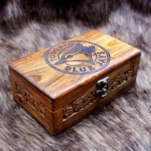 Peut inclure: Une boîte en bois avec le logo des Blue Jays de Toronto gravé sur le couvercle. La boîte rectangulaire a un couvercle à charnière et un fermoir en métal. Le bois est de couleur marron chaud, et la boîte est ornée de motifs floraux sculptés.