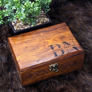 Peut inclure: Une boîte en bois rectangulaire avec un couvercle à charnière et un fermoir en métal. La boîte est gravée des initiales "DNT" et du nom "BAILEY NEWTON". Le bois est brun chaud avec un grain visible.