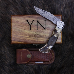 Peut inclure: Un couteau pliant personnalisé avec une lame à motifs et un manche en corne de cerf. Il est présenté avec un coffret en bois gravé des initiales "YN" et de l'inscription "Your Name". Un étui en cuir est également inclus.